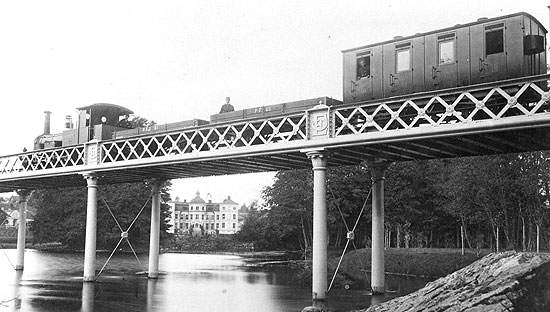 Bilden visar lok nummer 1 "Finspong" med blandat tåg på den vackra bron över Finspångsån. Året är 1880 och i fonden syns Finspångs slott grundlagt av en son till Louis De Geer 1668