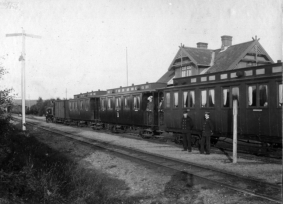 Ångloksdraget persontåg under uppehåll framför stationshuset. Personvagnarna, två tvåaxliga och en Boggivagn, samtliga med lanternintak . 