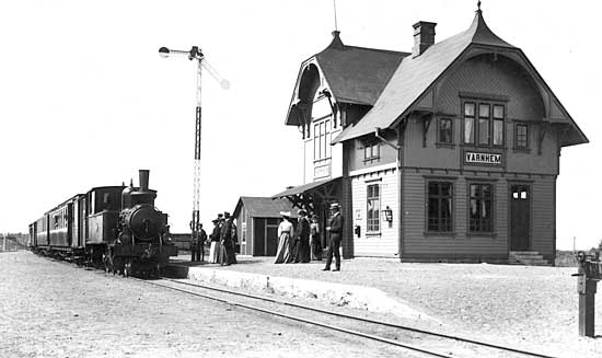 SAJ lok nummer 2 "VARNHEM" med persontåg ankommer Varnhem. Året är 1904 och järnvägen har nyss öppnats för trafik. Den uniformerade mannen till höger om semaformasten är stationens förste stationsinspektor, Otto Rosander